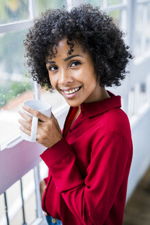 Porträt einer lächelnden Frau mit einer Tasse Kaffee am Fenster zu Hause - GIOF05541