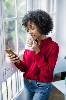 Frau mit Tasse Kaffee und Mobiltelefon am Fenster stehend zu Hause - GIOF05542