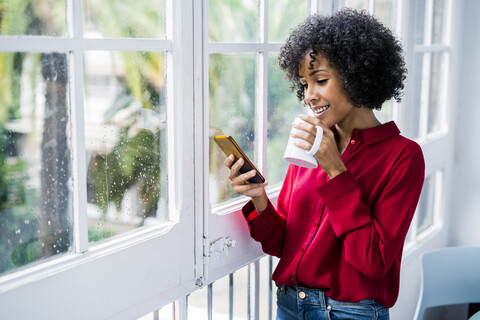 Smiling woman with cup of coffee and cell phone standing at the window at home stock photo