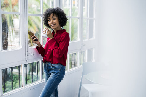 Porträt einer lächelnden Frau mit einer Tasse Kaffee und einem Handy, die zu Hause am Fenster steht - GIOF05544
