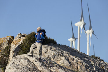 Spanien, Andalusien, Tarifa, Mann beim Wandern auf einem Felsen stehend mit Windrädern im Hintergrund - KBF00416
