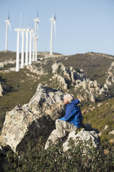 Spanien, Andalusien, Tarifa, Mann beim Wandern auf einem Felsen sitzend mit Windrädern im Hintergrund - KBF00419