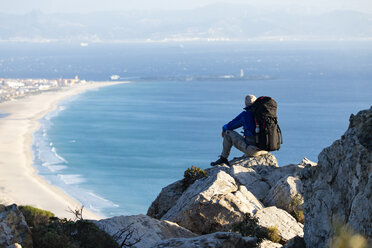 Spanien, Andalusien, Tarifa, Mann beim Wandern an der Küste, auf einem Felsen sitzend und die Aussicht betrachtend - KBF00427