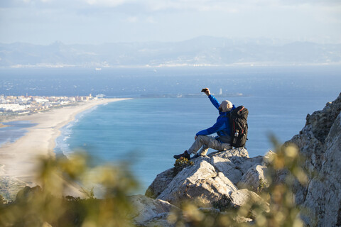Spanien, Andalusien, Tarifa, Mann beim Wandern an der Küste auf einem Felsen sitzend und ein Selfie machend, lizenzfreies Stockfoto