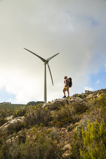 Spain, Andalusia, Tarifa, man on a hiking trip standing on rock with wind turbine in background - KBF00435