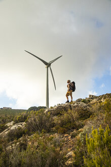 Spanien, Andalusien, Tarifa, Mann beim Wandern auf einem Felsen stehend mit Windrad im Hintergrund - KBF00435