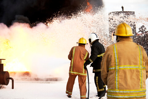 Feuerwehrausbildung, ein Team von Feuerwehrleuten sprüht Löschschaum auf ein Feuer in einer Übungsanlage, Rückansicht, lizenzfreies Stockfoto