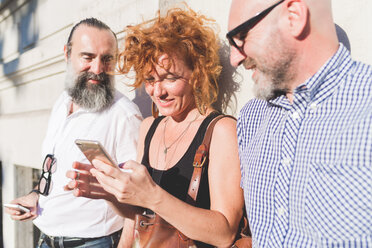 Mid adult woman and male friends leaning against wall looking at smartphone - CUF48237