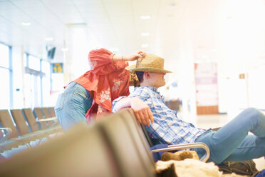 Junger Mann sitzt am Flughafen, junge Frau setzt ihren Hut auf seinen Kopf - ISF20123