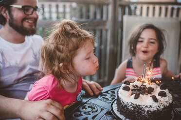 Weibliches Kleinkind mit Schwester und Vater beim Ausblasen von Wunderkerzen auf der Festtorte am Terrassentisch - ISF20186