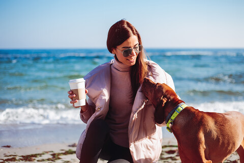 Stilvolle mittelgroße erwachsene Frau hockt am Strand und streichelt ihren Hund, Odessa, Odeska Oblast, Ukraine, lizenzfreies Stockfoto