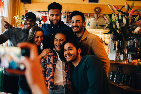 Young woman photographing cheerful multi-ethnic friends standing at restaurant during brunch - MASF10861