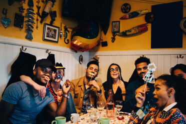 Portrait of cheerful young multi-ethnic friends holding props while sitting at restaurant during dinner party - MASF10866