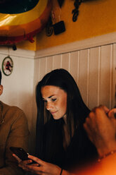 Young woman using smart phone while sitting amidst multi-ethnic male friends in restaurant - MASF10870