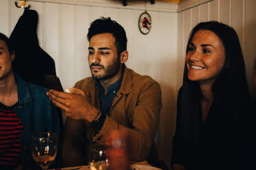 Junger Mann, der sein Smartphone benutzt, während er inmitten von Freundinnen in einem Restaurant beim Abendessen sitzt - MASF10874