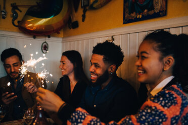 Happy multi-ethnic male and female friends holding burning sparklers in restaurant during dinner party - MASF10876