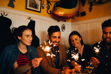 Happy young multi-ethnic male and female friends holding burning sparklers in restaurant during dinner party - MASF10880