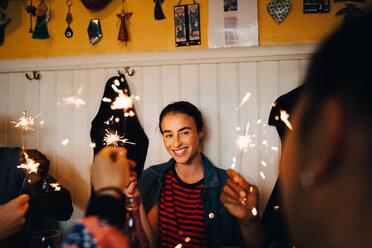 Smiling young multi-ethnic friends holding burning sparklers while enjoying in restaurant during dinner party - MASF10883