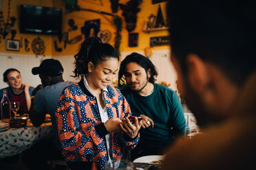 Smiling young woman sharing smart phone with male friend while sitting at restaurant during dinner party - MASF10886