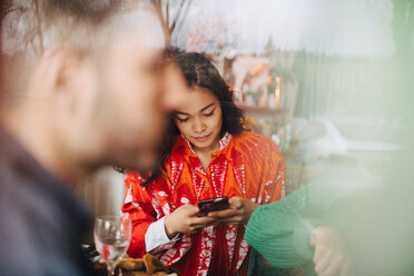 Junge Frau benutzt ihr Smartphone, während sie mit Freunden in einem Restaurant sitzt, gesehen durch ein Glasfenster - MASF10887