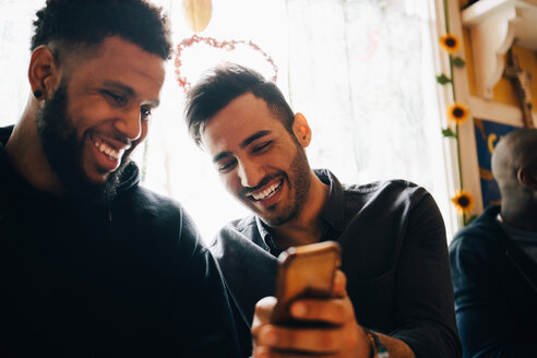 Fröhlicher junger Mann zeigt einem Freund sein Smartphone, während er während einer Brunchparty im Restaurant am Fenster sitzt - MASF10892
