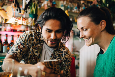 Smiling young man sharing smart phone with woman sitting in restaurant during brunch party - MASF10893