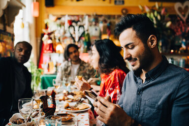 Lächelnder junger Mann, der eine SMS auf seinem Smartphone schreibt, während er mit Freunden an einem Tisch im Restaurant sitzt - MASF10897