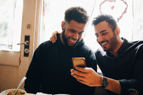 Lächelnde junge Männer, die sich ein Smartphone teilen, während sie während einer Brunch-Party im Restaurant am Fenster sitzen - MASF10899