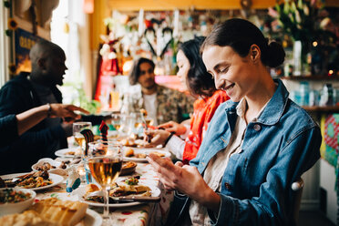 Lächelnde junge Frau, die ein Smartphone benutzt, während sie mit Freunden an einem Tisch im Restaurant sitzt - MASF10900