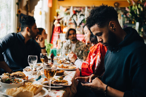 Junger Mann schreibt Textnachrichten über sein Smartphone, während er mit Freunden bei einer Brunchparty im Restaurant sitzt, lizenzfreies Stockfoto