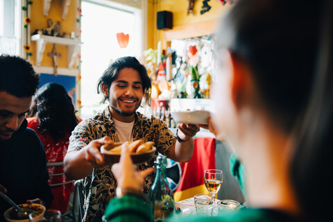 Lächelnder junger Mann, der einer Frau Essen gibt, während er mit einem Freund an einem Tisch im Restaurant während einer Brunchparty sitzt, lizenzfreies Stockfoto