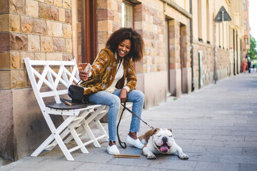 Happy woman sitting with dog taking selfie on mobile phone at sidewalk in city - MASF10920