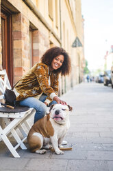 Smiling woman sitting with dog on sidewalk in city - MASF10926