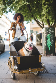 Lächelnde Frau auf Fahrradkarren mit Bulldogge auf einer gepflasterten Straße - MASF10928