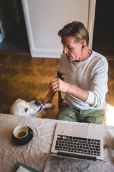 High angle view of senior man playing with dog while working on laptop from home - MASF10933