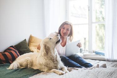 Loving senior woman touching dog while holding laptop on bed at home - MASF10940