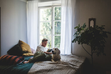 Full length of retired man reading book while resting with dog on bed at home - MASF10942