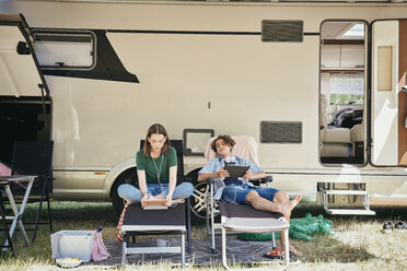 Geschwister im Teenageralter sehen sich einen Film auf einem digitalen Tablet an, während sie auf Klappstühlen vor dem Wohnmobil auf dem Campingplatz sitzen - MASF10962