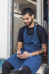 Junger männlicher Unternehmer schreibt Textnachrichten, während er am Eingang eines Food Trucks sitzt - MASF11013