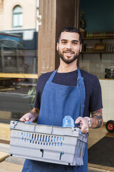 Porträt eines lächelnden jungen männlichen Besitzers, der eine Kiste gegen ein Restaurant trägt - MASF11022