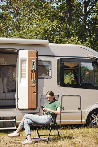 Teenager-Mädchen benutzt Mobiltelefon, während sie auf einem Klappstuhl am Wohnmobil sitzt, lizenzfreies Stockfoto