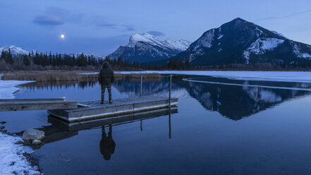 Frau an einem ruhigen Winterseesteg mit Blick auf den Vollmond über den Bergen, Banff, Alberta, Kanada - FSIF03717