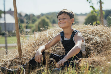 Porträt eines selbstbewussten, lächelnden Jungen in Latzhose, der auf einem sonnigen Bauernhof Heu kaut - FSIF03723