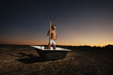 Porträt selbstbewusster Junge in Unterwäsche, der eine Mistgabel in einer Badewanne in einem ländlichen Gebiet hält - FSIF03733