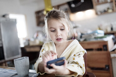 Porträt eines kleinen Mädchens, das am Frühstückstisch in der Küche sitzt und ein Smartphone benutzt - KMKF00715