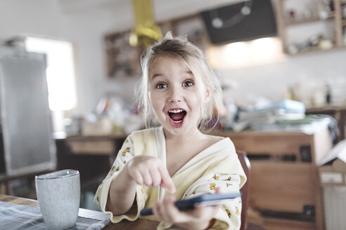 Portrait of excited little girl in the kitchen pointing at smartphone - KMKF00716