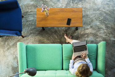 Draufsicht auf eine Geschäftsfrau, die auf einer türkisfarbenen Couch im Coworking Space am Laptop arbeitet - SBOF01544