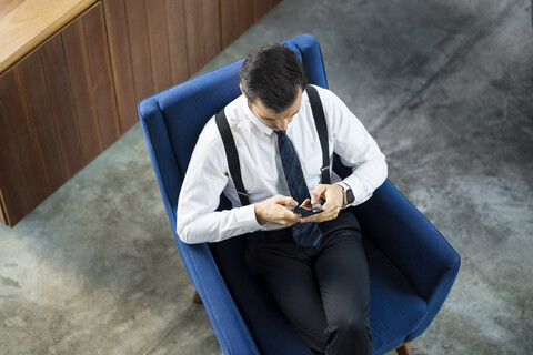 Draufsicht auf einen Geschäftsmann, der in einem blauen Sessel sitzt und ein Smartphone benutzt, lizenzfreies Stockfoto