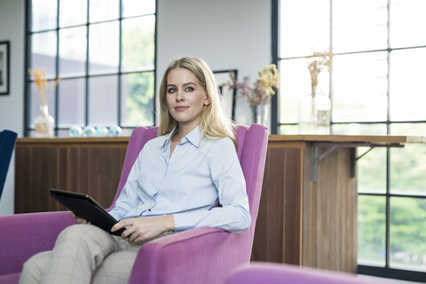 Content blond businesswoman sitting on violet armchair with tablet stock photo