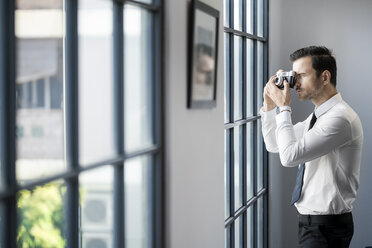 Geschäftsmann fotografiert mit einer Retro-Kamera vor einem großen Bürofenster - SBOF01575